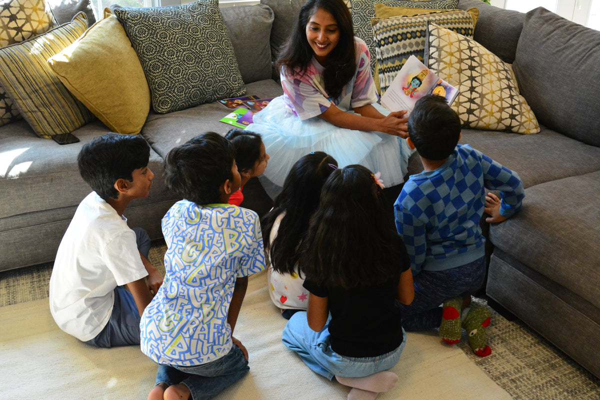 A woman sitting on a gray sectional sofa reads a book to a group of children who are kneeling on a light-colored rug in a living room.